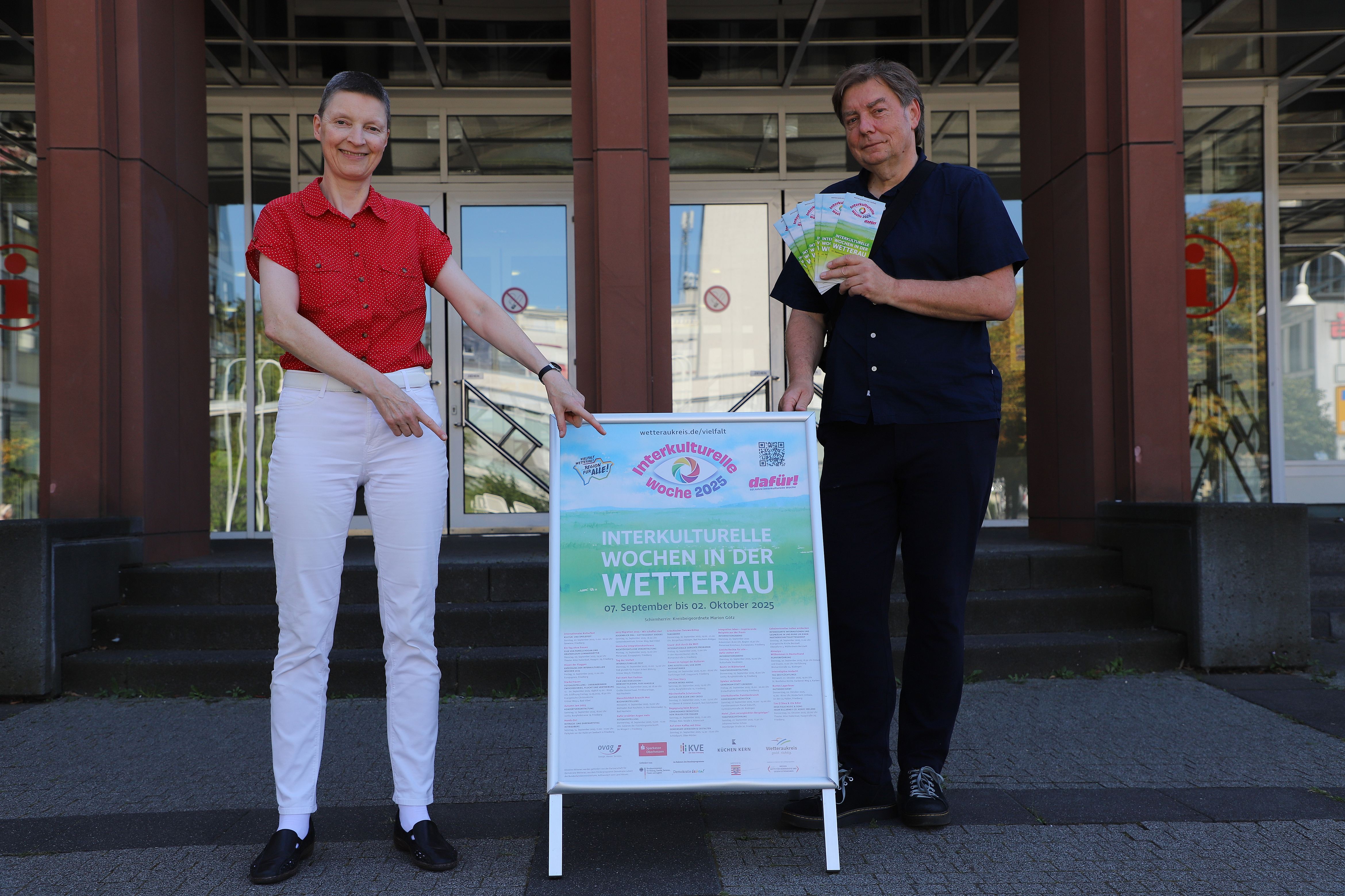 Eine Frau und ein Mann stehen vor einem Gebäude. Zwischen beiden befindet sich ein Plakat, auf das die Frau zeigt. Der Mann hält mehrere Flyer in der Hand.