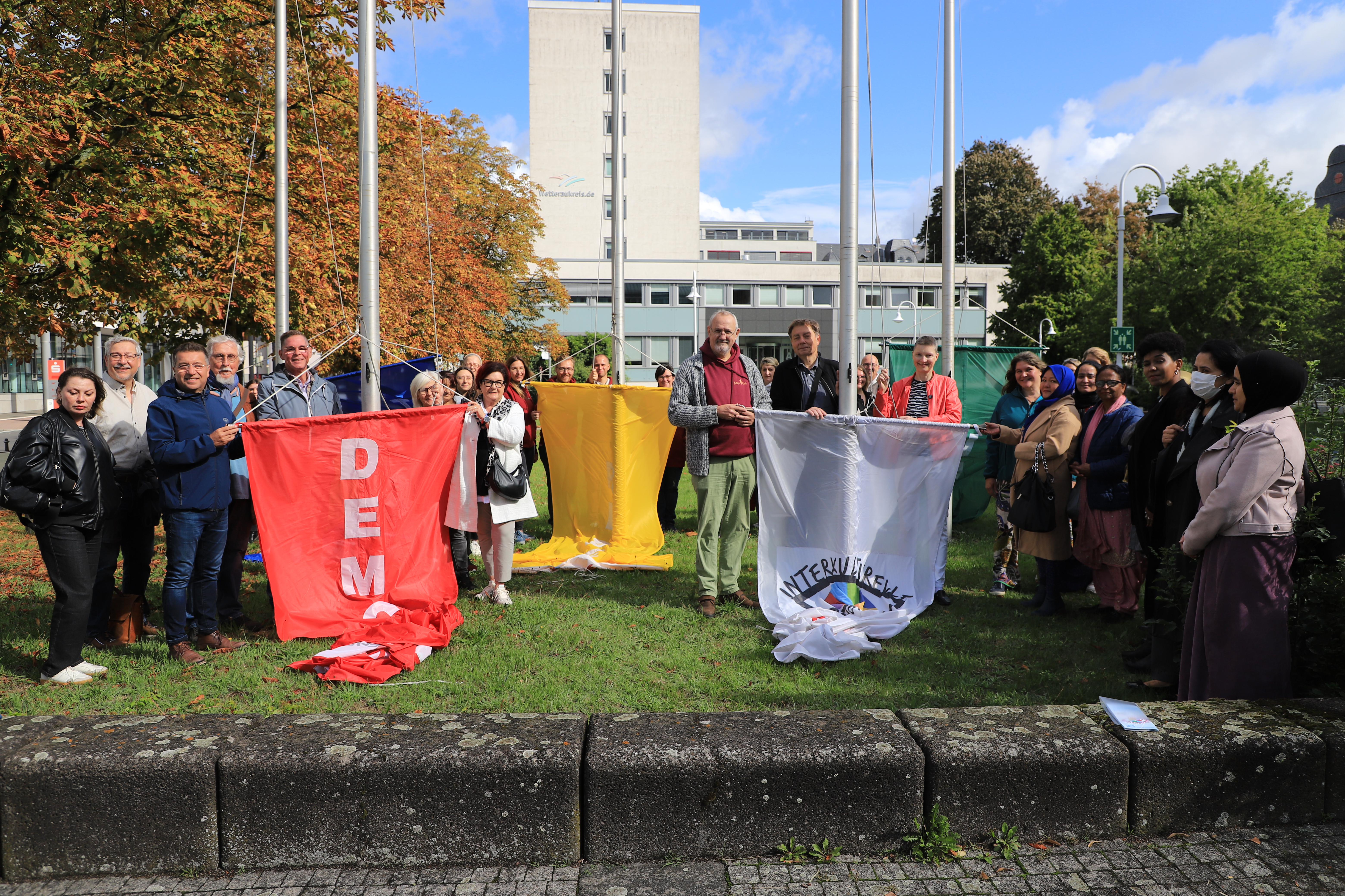 Auf einem Platz mit einem mehrstöckigen Bürogebäude im Hintergrund steht eine Gruppe von Menschen an mehreren Fahnenmasten. Sie bereiten sich darauf vor, mehrere Fahnen an den Masten emporzuziehen.