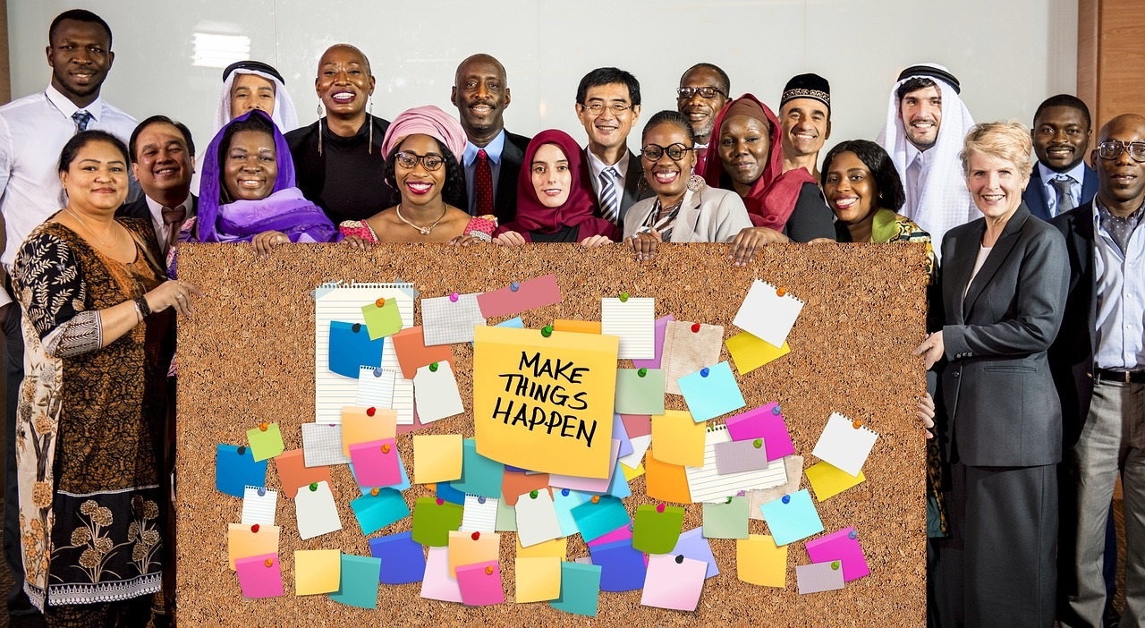 Eine Gruppe von erwachsenen Personen mit unterschiedlichen Hautfarben und Aussehen halten ein großes Plakat. Auf dem Plakat gibt es viele kleine bunte Zettel und ein großes Zettel mit dem Text "Make things happen". 