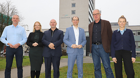Zwei Frauen und vier Männer stehen gemeinsam im Freien auf einem Platz. Im Hintergrund sind ein Hochhaus und weitere Gebäude zu erkennen.