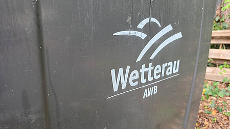 Eine Mülltonne mit der Aufschrift AWB und einem Logo steht im Freien.