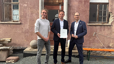 Drei Männer stehen vor der Fassade eines sanierungsbedürftigen Gebäudes. Einer der Männer hält eine Dokumentenmappe in Händen.