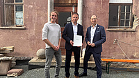 Drei Männer stehen vor der Fassade eines sanierungsbedürftigen Gebäudes. Einer der Männer hält eine Dokumentenmappe in Händen.