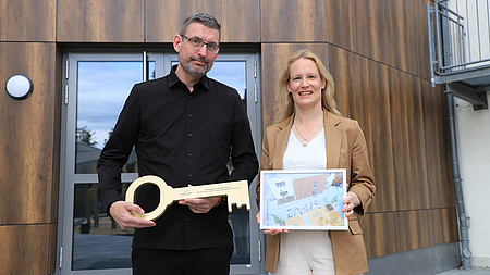 Eine Frau und ein Mann stehen vor der Eingangstür eines Gebäudes. Der Mann hält einen großen, goldenen Schlüssel in der Hand, die Frau ein gerahmtes Foto.