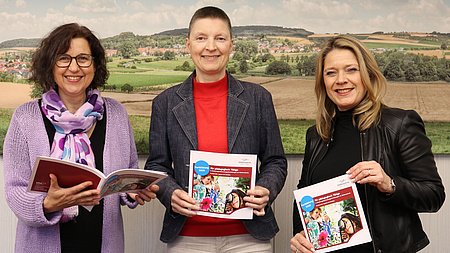 Andrea Rosenberger, Marion Götz und Ann-Karin Schild lächeln in die Kamera. Sie halten Broschüren in der Hand. Im Hintergrund ein Lanschaftsbild
