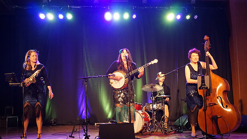 Vier musizierende Frauen stehen auf einer Bühne. Sie spielen Saxofon, Banjo, Schlagzeug und Kontrabass.