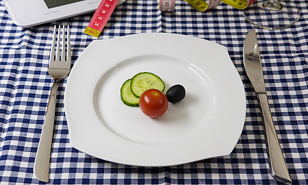 Auf einem Teller liegen eine Olive, eine kleine Tomate und zwei Gurkenscheiben. Daneben Messer und Gabel.