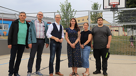 Vier Männer und zwei Frauen stehen auf einem Sportgelände, unter anderem mit einem Basketballkorb und einem Fußballtor, das vor einem Gebäude angelegt ist.