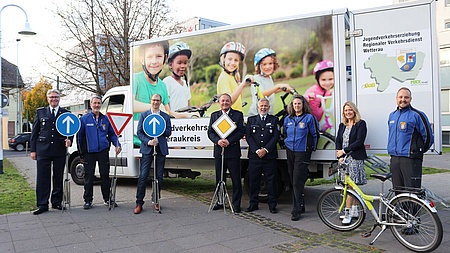 Eine Personengruppe. Vier von ihnen halten ein Verkehrsschild. Im Vordergrund ein Fahrrad, im Hintergrund ein Kleintransporter mit der Aufschrift Jugendverkehrserziehung Regionaler Verkehrsdienst Wetterau.