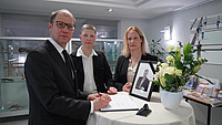 Drei Personen stehen an einem Stehtisch und trägt sich in ein Kondolenzbuch ein. Auf dem Tisch steht außerdem ein Foto und ein Blumengesteck.