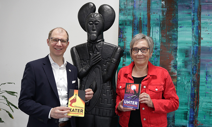 Ein Mann und eine Frau halten jeweils ein Buch in der Hand. Im Hintergrund eine Statue und ein buntes, abstraktes Gemälde.