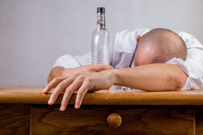 Mann liegt mit Kopf und verschränkten Armen auf Tisch, zwischen den Armen steht eine leere Flasche Alkohol, Quelle Pixabay, Michal Jarmoluk