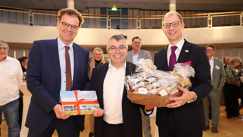 Drei Männer stehen vor dem Publikum des Neujahrsempfang. Übergeben wird ein Präsentkorb und ein Buch.