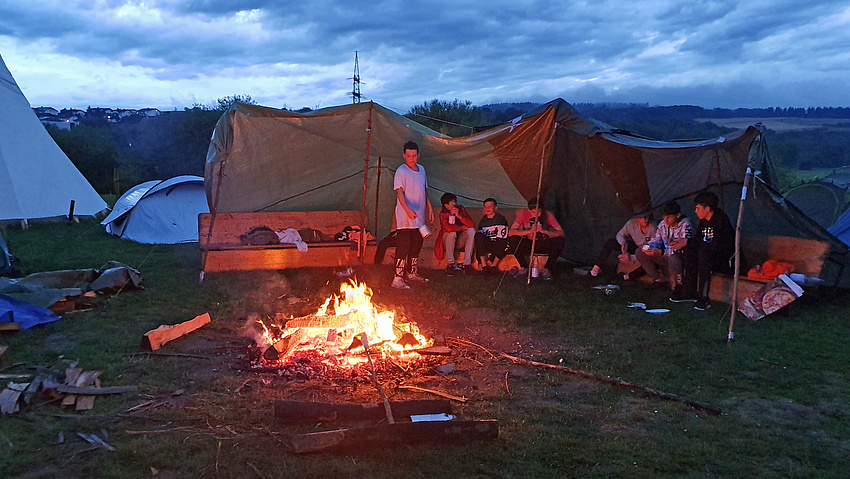 Mehrere Zelte um ein brennendes Lagerfeuer. Vor den offenen Zelten sitzen und stehen Kinder.