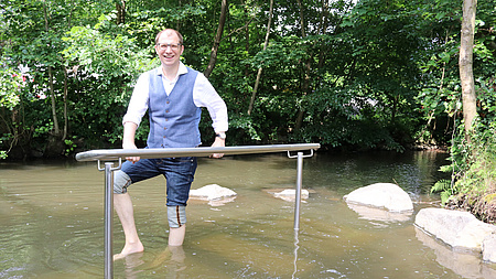 Landrat Weckler steht Barfuß im Bach, vor im eine Haltestange