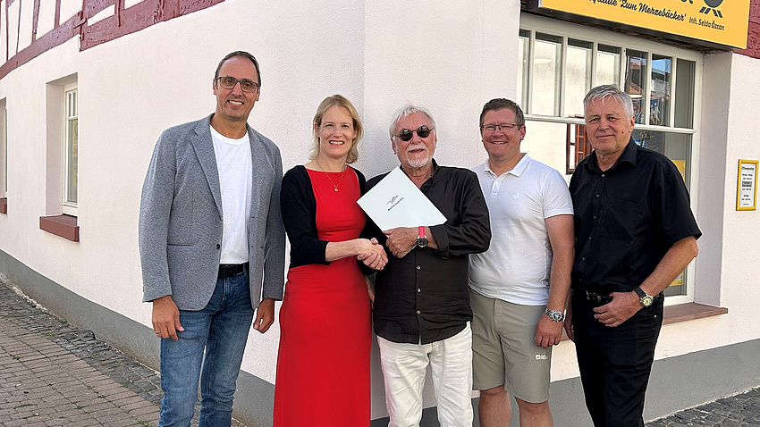 Fünf Personen stehen bei der Übergabe des Förderbscheids vor einem sanierten Fachwerkhaus. Das Haus ist weiß mit roten Balken.