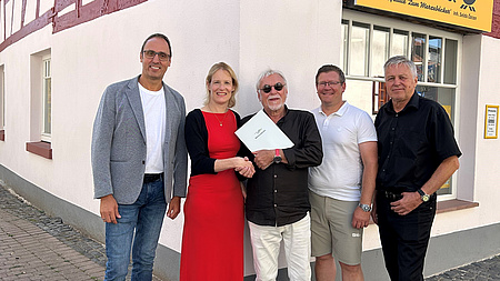 Fünf Personen stehen bei der Übergabe des Förderbscheids vor einem sanierten Fachwerkhaus. Das Haus ist weiß mit roten Balken.