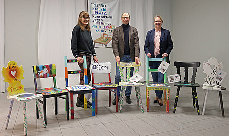 Ein Mann und zwei Frauen stehen hinter acht bunt angemalten Stühlen.