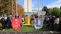 Auf einem Platz mit einem mehrstöckigen Bürogebäude im Hintergrund steht eine Gruppe von Menschen an mehreren Fahnenmasten. Sie bereiten sich darauf vor, mehrere Fahnen an den Masten emporzuziehen.