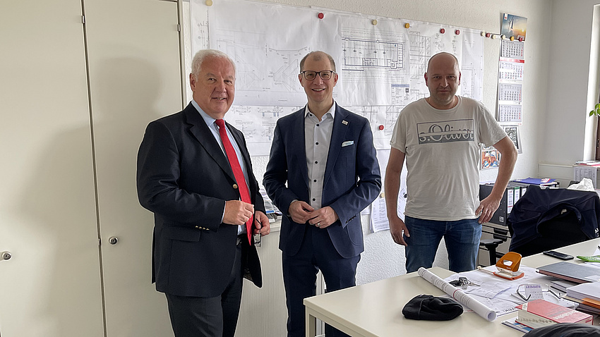 Drei Männer stehen in einem Raum und lächeln in die Kamera. An der Wand hinter ihnen hängen Baupläne.