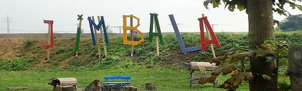 Auf einer Wiese sind auf Pfählern einzeln die verschiedenfarbenen Buchstaben des Wortes Jimbala auf Holz aufgesteckt.