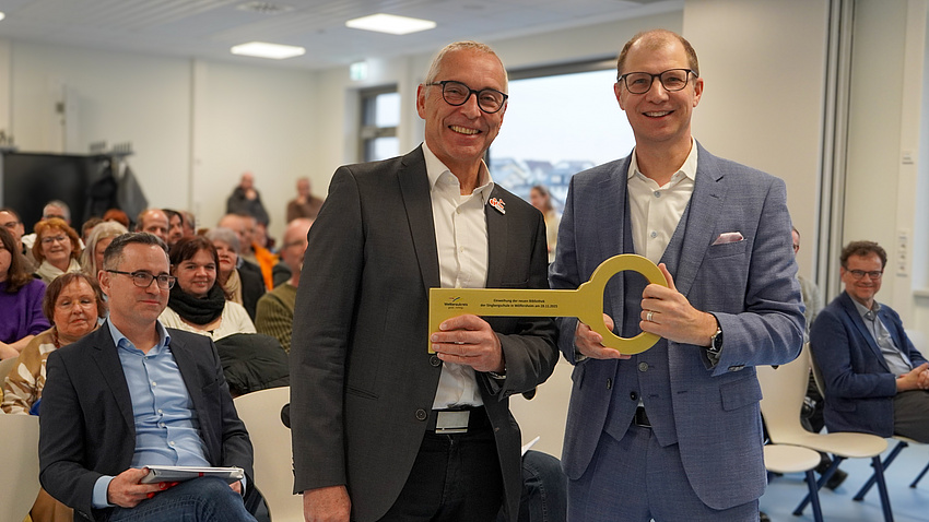 In einem Saal stehen zwei Männer vor einer größeren Gruppe auf Stühlen sitzender Personen. Beide Männer halten gemeinsam die Nachbildung eines goldenen Schlüssels in Händen.