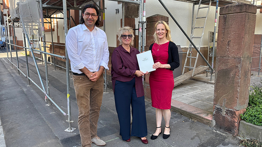 Zwei Frauen und ein Mann stehen auf einem Bürgersteig vor einem Baugerüst an einer Hauswand. Die beiden Frauen halten zusammen ein Dokument in Händen.