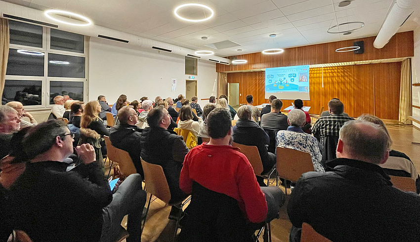 In einem Saal sitzen Menschen, sie sind von hinten zu sehen. Vorne wird eine Präsentation gezeigt.