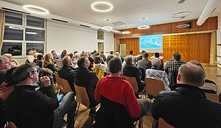 In einem Saal sitzen Menschen, sie sind von hinten zu sehen. Vorne wird eine Präsentation gezeigt.