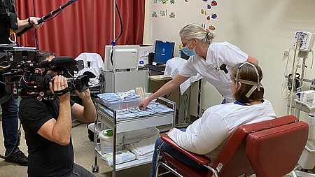 Die Patientin im Gespräch mit dem Arzt. Daneben ein Kameramann und ein Techniker, der ein Mikrofon hält.
