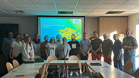 Mehrere Männer und Frauen stehen nebeneinander hinter Tischen, die in dder Form eines U aufgestellt sind.