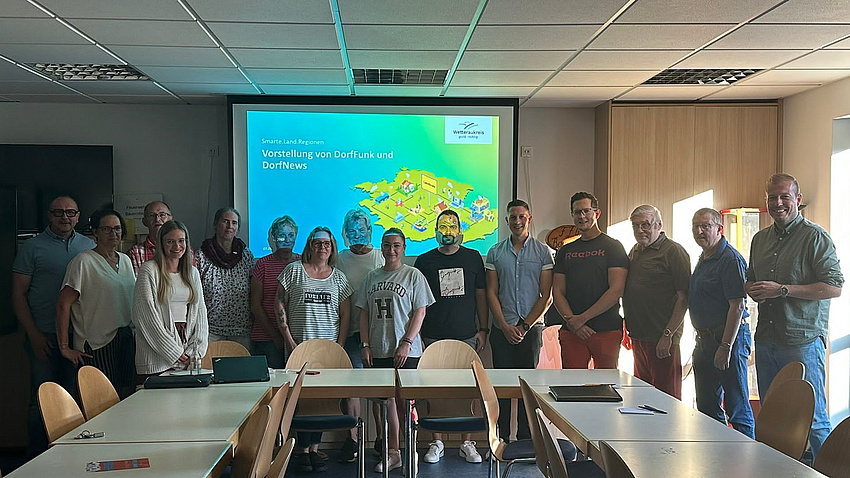 Mehrere Männer und Frauen stehen nebeneinander hinter Tischen, die in dder Form eines U aufgestellt sind.