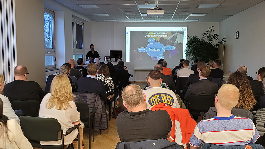 Mehrere Personen in einem größeren Raum hören einem Vortrag zu. Sie sitzen in Stuhlreihen und sind von hinten zu sehen. Vorne referiert ein Mann.