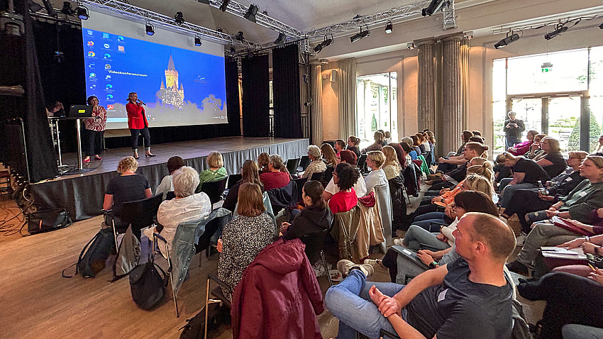 Menschen in einem Saal mit Reihenbestuhlung. Vorne eine Bühne, auf der zwei Frauen zum Publikum sprechen. Hinter ihnen eine Leinwand mit einer Präsentation.