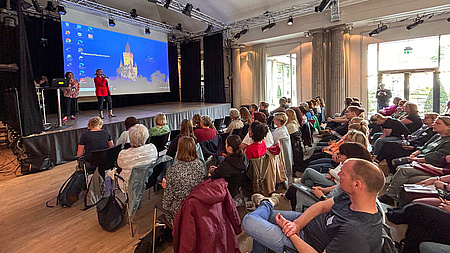Menschen in einem Saal mit Reihenbestuhlung. Vorne eine Bühne, auf der zwei Frauen zum Publikum sprechen. Hinter ihnen eine Leinwand mit einer Präsentation.