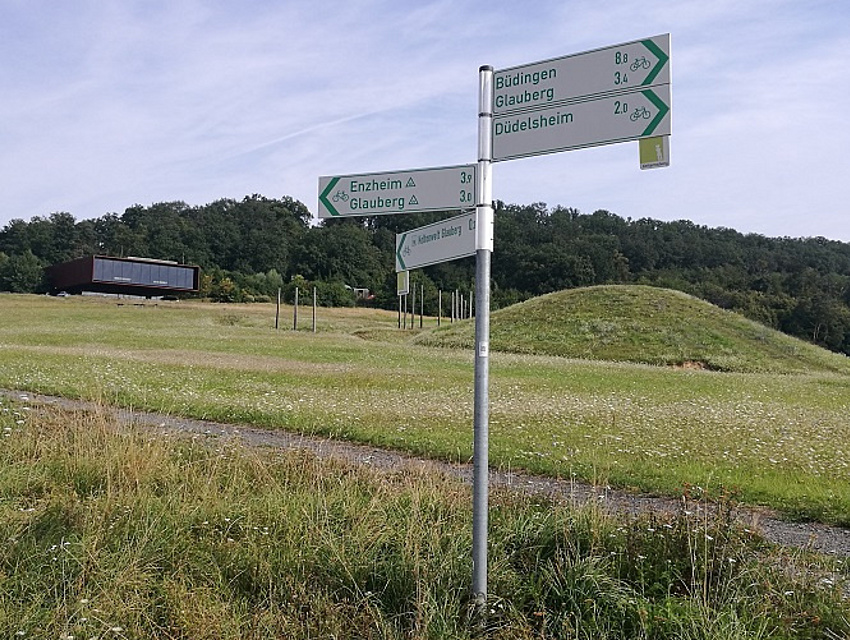 : Neue Radwegebeschilderung im Vordergrund des Bildes zu sehen, im Hintergrund ist der Glauberg und die Keltenwelt zu sehen.