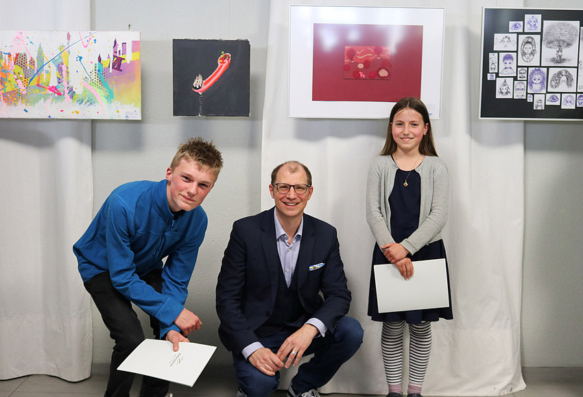 Ein Mann in der Hocke, links daneben ein Junge in gebückter Haltung. Rechts steht ein Mädchen. Die Kinder halten Urkunden in der Hand. Im Hintergrund hängen Bilder an der Wand.