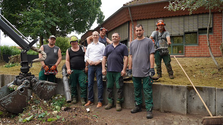 Mehrere Männer stehen vor einem Gebäude auf der Wiese und haben Gerätschaften zur Gartenarbeit in den Händen.