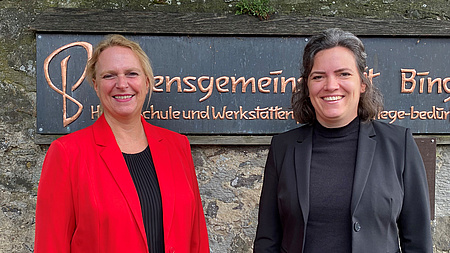 Die beiden Frauen stehen vor einer Kupfertafel mit dem Schriftzug Lebensgemeinschaft Bingenheim.