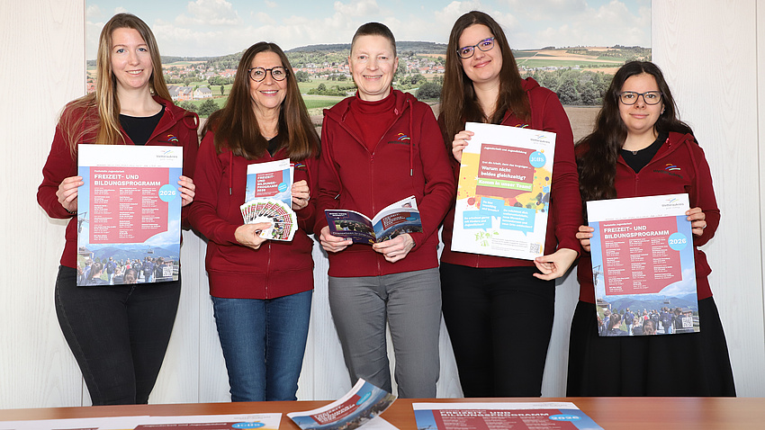 Fünf Frauen stehen in einem Raum vor einem Tisch. Die Frauen halten Broschüren und Plakate in Händen, die auch noch einmal auf dem Tisch liegen. Im Hintergrund ist ein Landschaftsfoto zu sehen.