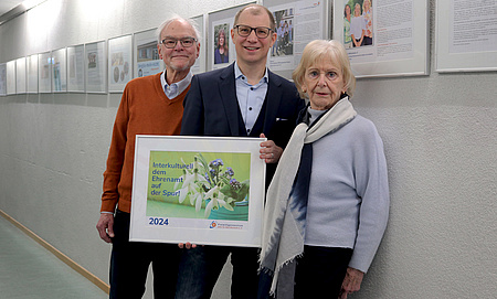 Eine Frau und zwei Männer stehen nebeneinander. Der Mann in der Mitte hält ein Bild mit einem Plakat in der Hand. Auf diesem steht: Interkulturell dem Ehrenamt auf der Spur. An der Wand im Hintergrund die weiteren Bilder der Ausstellung.