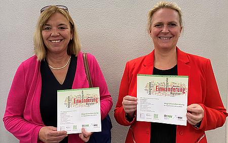 Zwei Frauen stehen nebeneinander. Jede von ihnen hat ein Informationsfaltblatt in der Hand