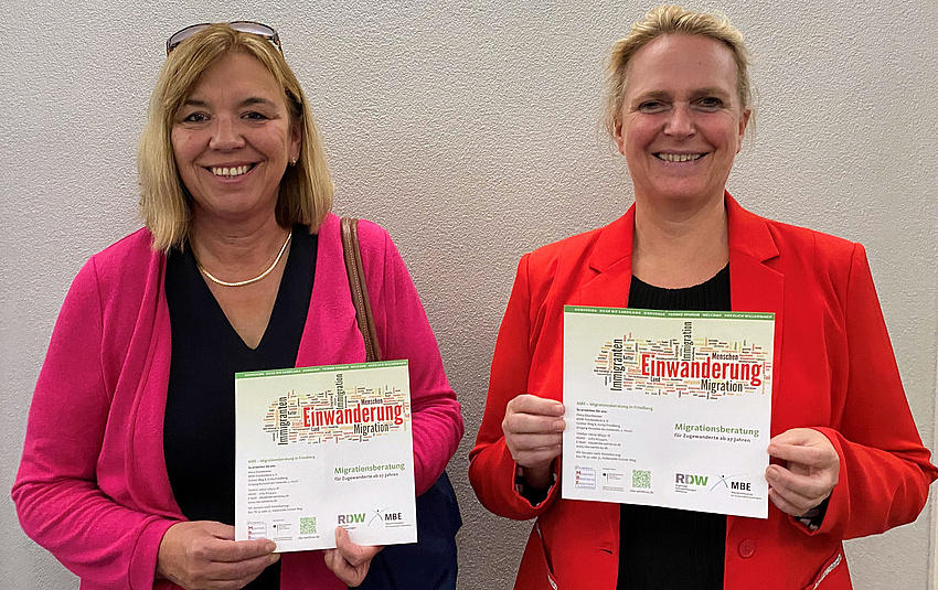 Zwei Frauen stehen nebeneinander. Jede von ihnen hat ein Informationsfaltblatt in der Hand