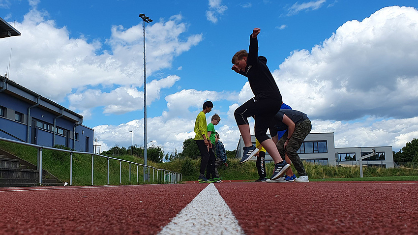 Jugendliche springen auf der Tartanbahn eines Sportplatzes von einer Linie zur anderen.