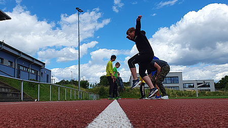 Jugendliche springen auf der Tartanbahn eines Sportplatzes von einer Linie zur anderen.