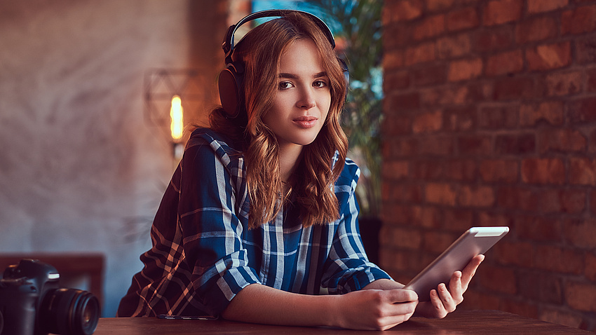 Ein Mädchen mit Kopfhörern auf dem Kopf und Handy in der Hand sitzend am Tisch