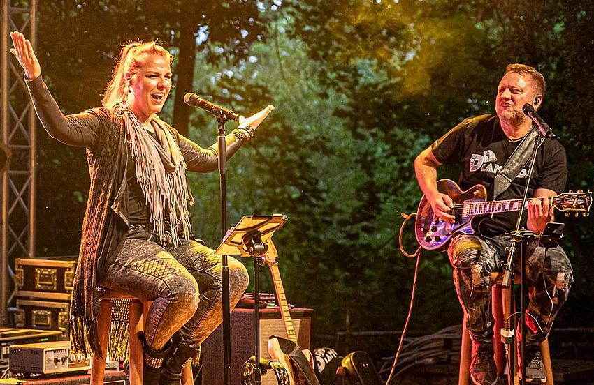 Ein Mann und eine Frau auf einer Bühne. Beide sitzen auf Barhockern. Der Mann spielt Gitarre, die Frau hat die Arme ausgebreitet.