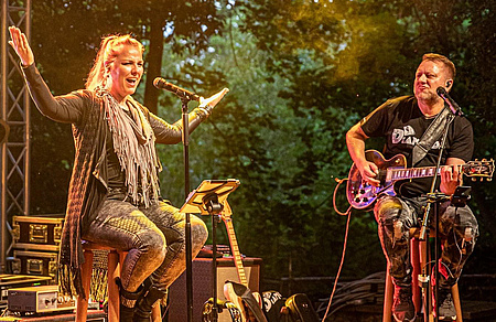 Ein Mann und eine Frau auf einer Bühne. Beide sitzen auf Barhockern. Der Mann spielt Gitarre, die Frau hat die Arme ausgebreitet.