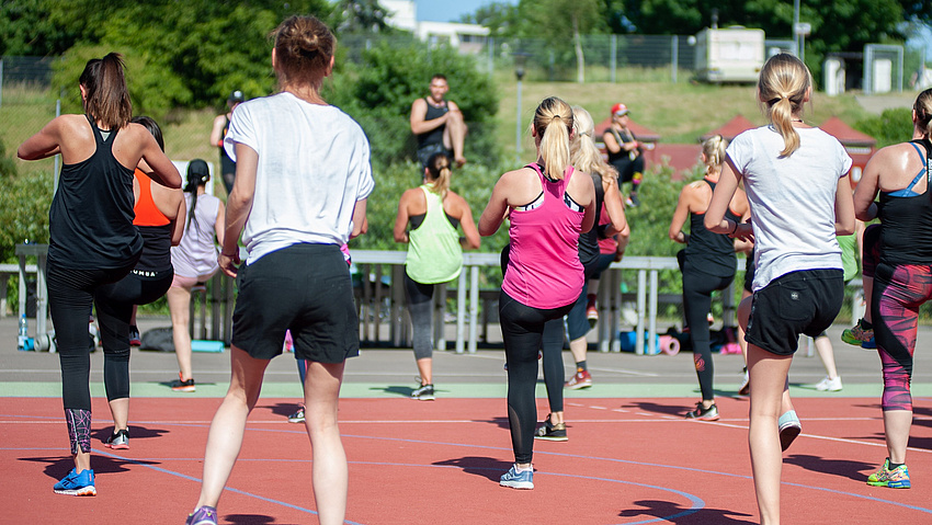Mehrere Frauen und Männer stehen auf der Tartanbahn eines Sportplatzes und absolvieren gemeinsam eine sportliche Übung.
