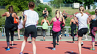 Mehrere Frauen und Männer stehen auf der Tartanbahn eines Sportplatzes und absolvieren gemeinsam eine sportliche Übung.